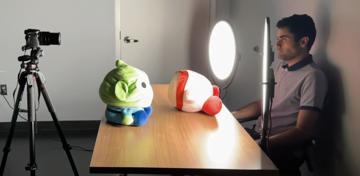 Bild im Fotolabor, Mann schaut in Kamera, auf dem Tisch vor ihm liegen zwei Plüschtiere, die Lampen leuchten die Szene hell aus.